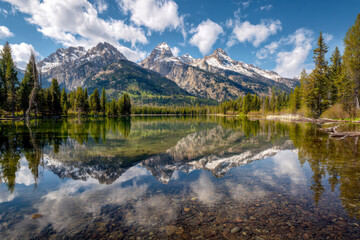 Stunning mountain range with snow-capped peaks reflected in a calm lake surrounded by lush green forest under a vibrant blue sky with fluffy clouds on a sunny day