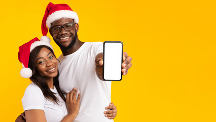 Great Application. African American Couple Showing Smartphone Empty Screen Wearing Santa Hats Advertising Christmas Offer Or Message Standing Over Yellow Background. Selective Focus On Phone