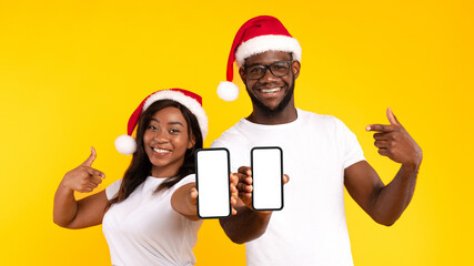 Black Couple Showing Phones Blank Screens Pointing Fingers And Gesturing Thumbs Up Approving Application Wearing Santa Hats Standing On Yellow Background. Christmas Offer For Smartphone Users