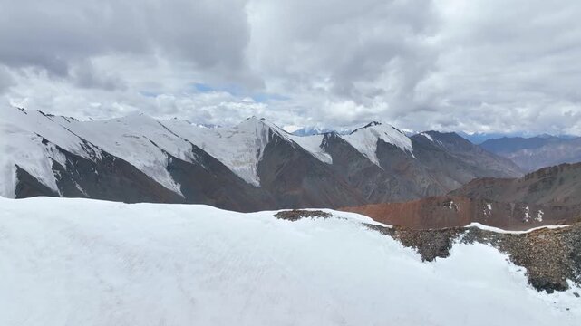 A dramatic panoramic view over a foreground of snow-covered high mountain terrain, facing an extensive, rugged range of peaks completely covered in permanent glaciers and snowfields under a cloudy sky