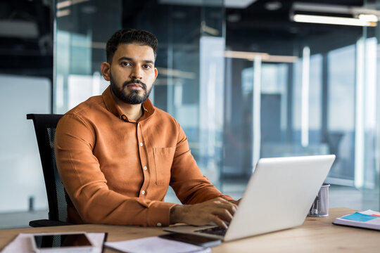 Indian business executive working on a laptop at his desk in a contemporary glass-walled office, focusing on his tasks and representing professionalism and corporate productivity