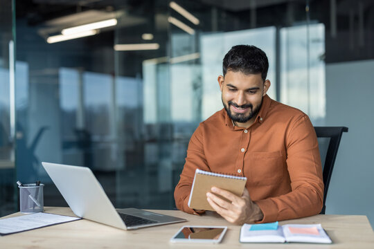 Young smiling indian businessman sitting at a desk in a modern office, concentrating on taking notes in a notebook while working with a laptop in a productive workspace - Powered by Adobe