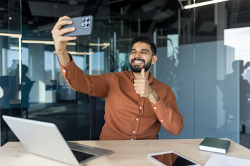 Young businessman sitting at his office desk smiling and giving a thumbs up while taking a selfie with his smartphone, conveying success, positivity and professional confidence