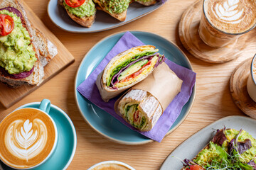 Fresh and healthy breakfast spread featuring vibrant avocado toast, colorful vegetable wraps, and expertly crafted lattes with detailed leaf art on wooden table