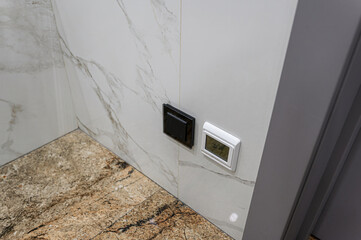 A close-up of a wall featuring a black light switch and a white thermostat with a digital display,...