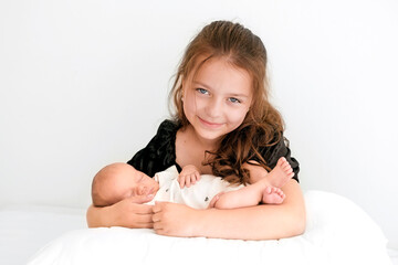 A cute beautiful girl holds her newborn baby brother in arms while sitting in bed. Love and help in caring for younger children