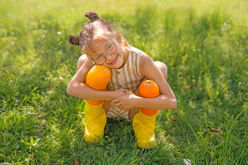 A cute, cheerful girl in rubber boots hugs two large yellow squash. The harvest season in the vegetable garden