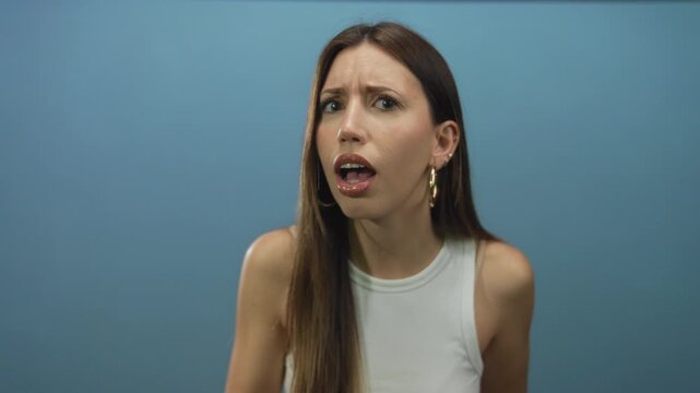 Hispanic woman wearing white tank top points finger and holds palm up with bare arm in blue studio with serious gaze; confidence assertion focus.