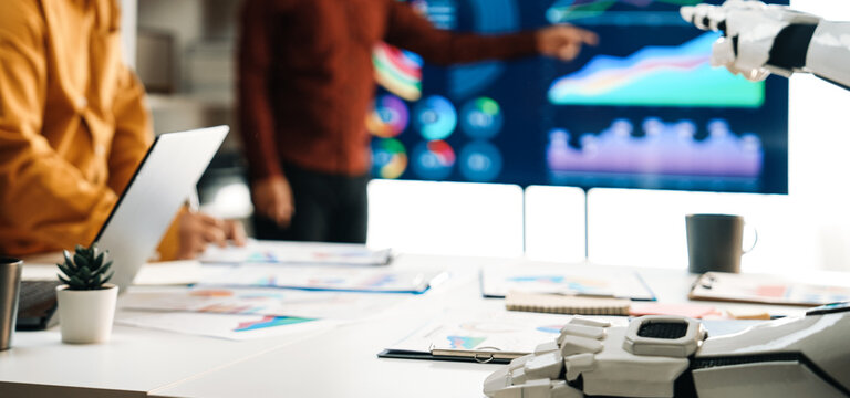 A dynamic business meeting showcases the integration of advanced technology with a robot hand assisting team members. Innovative visuals enhance strategic discussions.Noogenesis