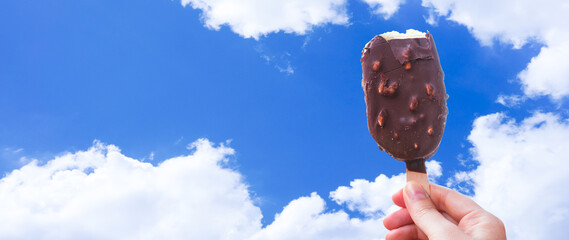 Chocolate covered ice cream bar with blue sky and cloud summer at the beach, during the holidays. Hand holds delicious chocolate and nuts almond ice cream bar on a stick.