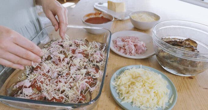 Woman prepares eggplant parmigiana, putting grated parmesan cheese on tomato sauce and fried slices of eggplant into lasagna pan