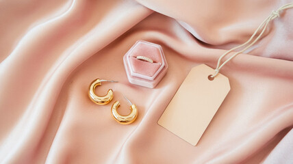 A gold ring in a pink velvet box sits beside crescent hoop earrings on silky pink satin, accented by a blank tag. Perfect for luxury gifts, fashion accessories, and jewelry showcases.