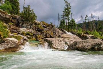 Crystal-clear mountain stream cascading over rocks in a forested landscape, surrounded by moss and trees under a dramatic cloudy sky. Pure nature in its raw beauty.