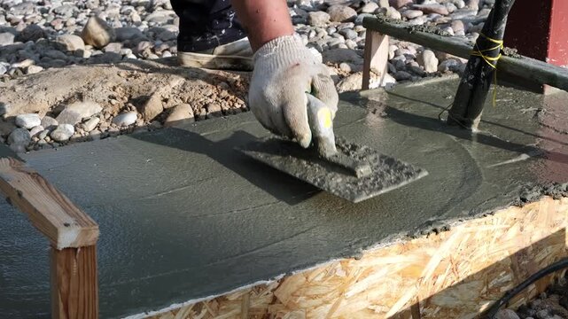 Construction worker smooths wet concrete with trowel in formwork on a building site. Smoothing a flat surface. Foundation work. Bundle of wires sticking out of a foundation for electrifying the gates.