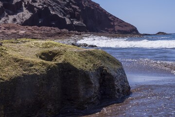 Blue ocean waves breaking on volcanic rocks along sandy beach. Idyllic coastal landscape with bright sky and sea horizon. Tenerife, Canary Islands, Spain.