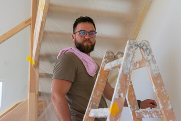 Bearded man with glasses renovating home interior and standing on ladder.