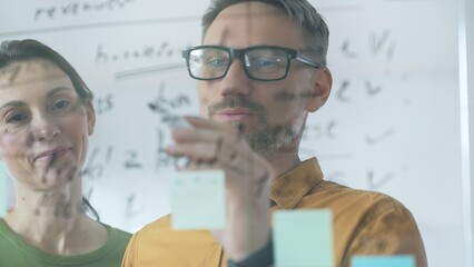 Professional team members brainstorming strategy, writing notes on transparent whiteboard with colorful sticky papers during collaborative meeting. Business people concept