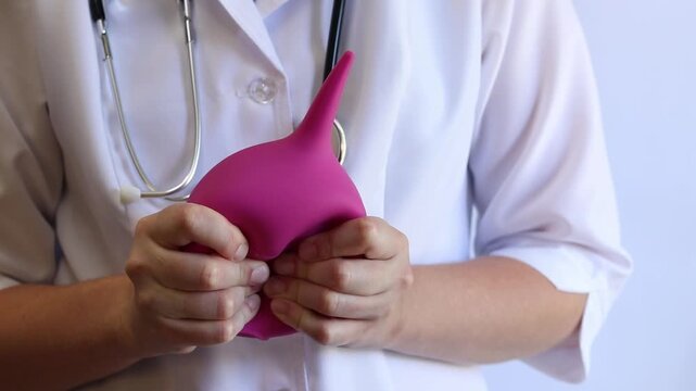 A female doctor in a white coat and with a stethoscope holds a large enema in her hands, close-up. A female doctor squeezes a pink rubber enema, checking for serviceability. Medical item. Nurse