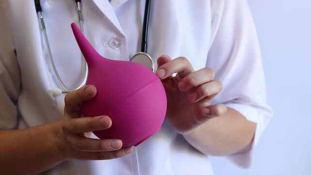 A female doctor in a white coat and with a stethoscope holds a large enema in her hands, close-up. A female doctor squeezes a pink rubber enema, checking for serviceability. Medical item. Nurse