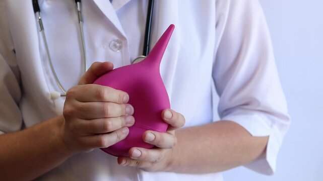 A female doctor in a white coat and with a stethoscope holds a large enema in her hands, close-up. A female doctor squeezes a pink rubber enema, checking for serviceability. Medical item. Nurse