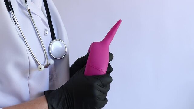 A female doctor in a white coat and with a stethoscope holds a large enema in her hands, close-up. A female doctor squeezes a pink rubber enema, checking for serviceability. Medical item. Nurse