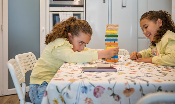 Concentrated twin sisters playing a wooden block stacking game at the kitchen table