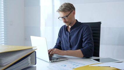 Obraz premium Professional businessman wearing glasses, intensely working on laptop, typing amid modern office environment with tablet and documents surrounding workspace. Business people concept