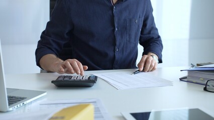 Businessman using a calculator and analyzing financial documents while preparing paperwork at an office desk. Audit and taxes, business concept