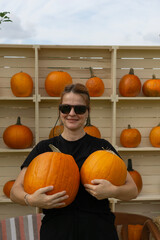 woman smiling holding two large orange pumpkins in front of a wooden shelf display outdoors with overcast sky, gazing and looking at camera, concept of farm market, seasonal decor, fall holiday