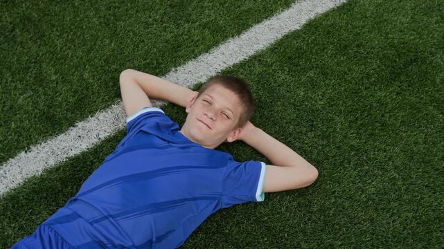 High angle shot of preteen Caucasian boy in blue football kit lying on fresh cut lawn with white touchlines. He smiling widely, making funny face