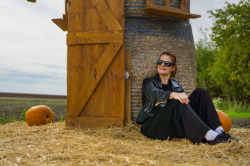 A woman in sunglasses sits on straw beside pumpkins near a wooden structure, enjoying an autumn day with a relaxed seasonal mood.