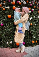 Mother holding child in front of decorated Christmas tree outdoors. Family enjoying Christmas season, winter holidays and New Year celebration together.
