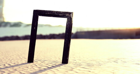Fototapeta premium A black rectangular frame is positioned upright on a sandy beach during sunset. The warm light creates dramatic long shadows, enhancing the tranquil atmosphere of the seaside.