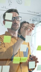 Business people team brainstorming, sharing strategy on glass whiteboard with colorful sticky notes, working collaboratively in bright office