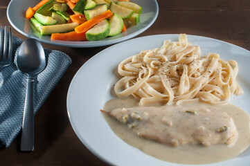 mushroom cream on a chicken breast with pasta on a white plate with vegetables and a gray napkin on one side on a wooden table, with cuttlery on the side