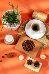 Cup of hot tasty coffee on wooden desk, coffee beans, candle and house plant in a beautiful cozy composition from above on orange background in sunlight. Americano, espresso and roasted coffee grain