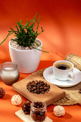Cup of hot tasty coffee on wooden desk, coffee beans, candle and house plant in a beautiful cozy composition from above on orange background in sunlight. Americano, espresso and roasted coffee grain