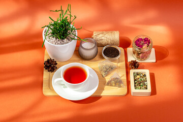 Mug of hot tasty tea on wooden desk, herbal tea leaves in glass bowls, candle and house plant in a beautiful cozy composition from above on orange background in sun light.