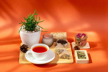 Mug of hot tasty tea on wooden desk, herbal tea leaves in glass bowls, candle and house plant in a beautiful cozy composition from above on orange background in sun light.