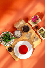 Mug of hot tasty tea on wooden desk, herbal tea leaves in glass bowls, candle and house plant in a beautiful cozy composition from above on orange background in sun light.