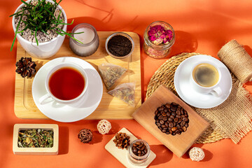 Cup of aromatic coffee and a mug of hot tasty tea on wooden desk, roasted coffee beans and herbal tea leaves in glass bowls, candle and house plant in cozy composition from above on orange background