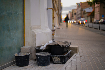 Construction buckets and renovation materials placed along building wall on urban sidewalk during evening representing city maintenance, repair and architectural restoration work