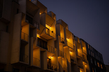 Apartment building glowing with balcony lights under dark evening sky, minimalist architectural...