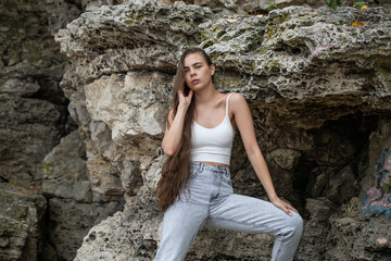 Beautiful woman in a white top and gray jeans poses on rocks in the mountains