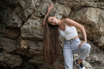 Beautiful woman in a white top and gray jeans poses on rocks in the mountains