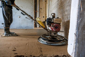 A worker operates a power trowel with a red engine on fresh concrete inside a building under construction, with exposed pipes and unfinished walls visible.