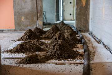 Construction site indoors showing soil piles on a white surface, a black pipe on the right, exposed concrete walls, and an open doorway in the background.