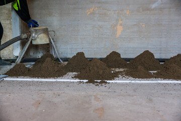 A construction site showing small piles of material on a flat surface. A worker in high visibility gear operates equipment near an unfinished wall.