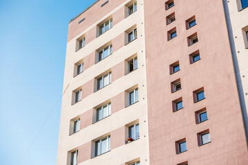 A multi story residential building with a pink and beige facade, rectangular and square windows, and a flower pot visible under a clear blue sky.