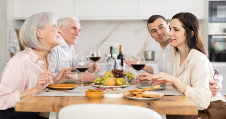 Mature couple are sitting at table, chatting sweetly, discussing and telling news, sharing plans with adult children. Senior woman and man celebrate housewarming party with middle-aged guests spouses